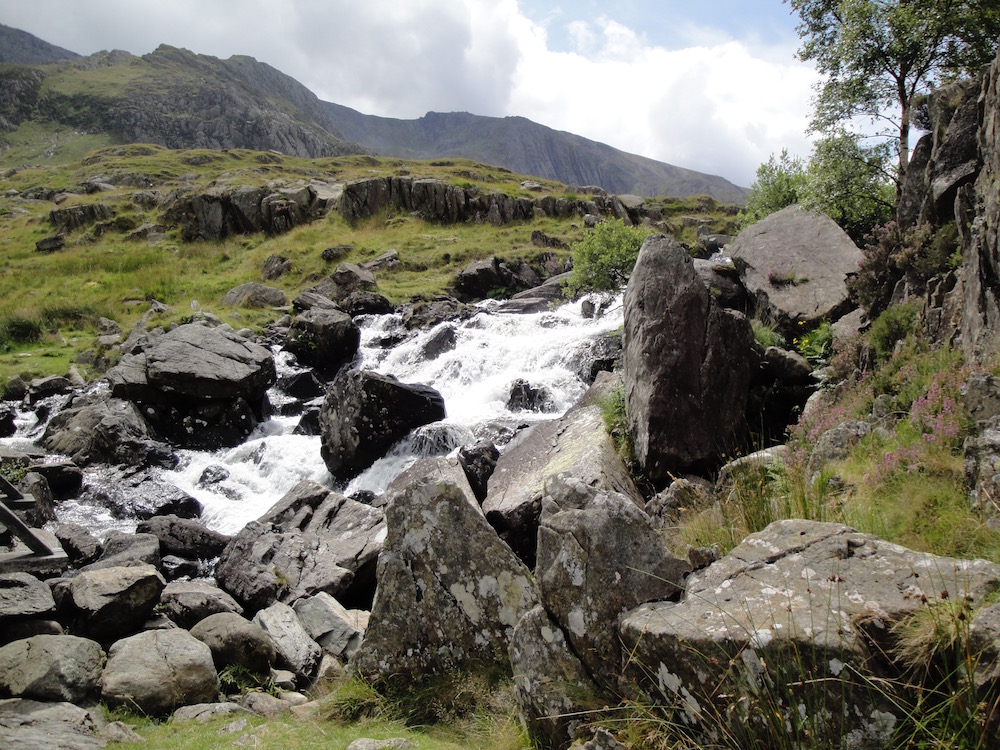 Berge im HIntergrund, Steinblöcke und ein kleiner Wasserfall im Bildvordergrund
